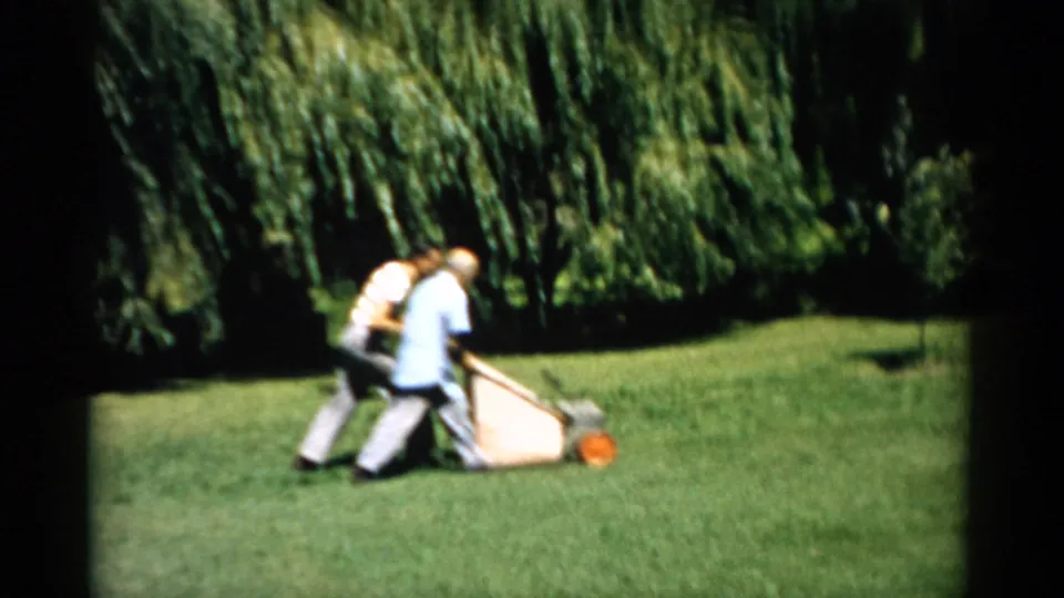 Two men in 1950s casual summer attire pushing a manual rotary mower across a sunlit lawn, Catskill, New York, circa 1957; weeping willows dominate the background.