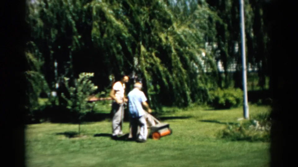 Two men operating a push-reel mower on a residential lawn, Catskill, New York, circa 1957; metal pole, weeping willows, and garden border plantings visible behind them.