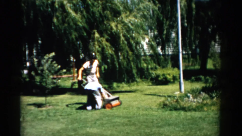Figure pushing a manual reel mower across a sunlit yard, Catskill, New York, circa 1957; weeping willow, metal pole, and garden plantings visible in background.