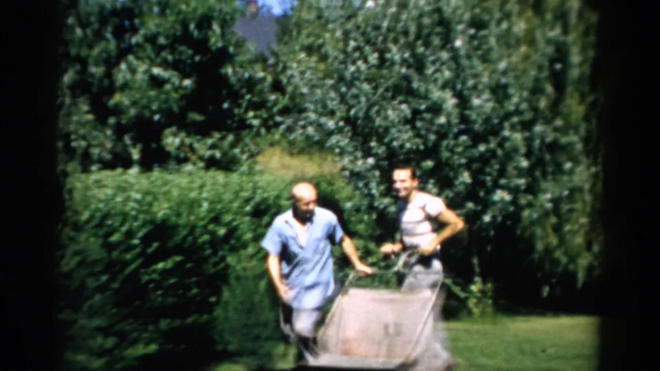 Two men push a vintage reel lawnmower with attached canvas grass catcher toward camera, green hedgerow behind; Catskill, NY, 1957.