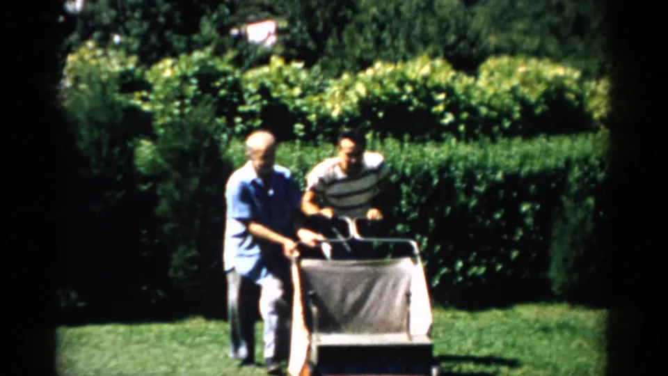 Two men beside a reel-type lawnmower with canvas grass catcher in a manicured garden, green hedges behind; Catskill, NY, 1957.