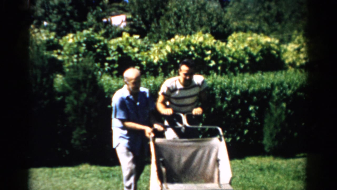 Two men, the older in a blue shirt and the younger in a striped t-shirt, push a wheeled metal cart across a sunny lawn in this 1950s Catskill, New York home movie.