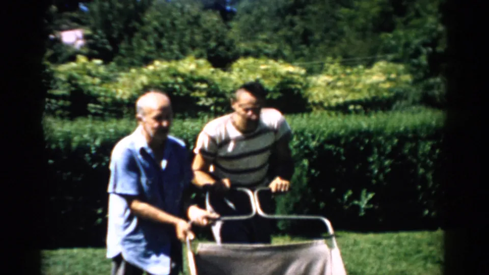 An older balding man in a blue shirt and a younger man in a striped t-shirt grip a metal cart together in a sunny backyard in this 1950s home movie from Catskill, New York.