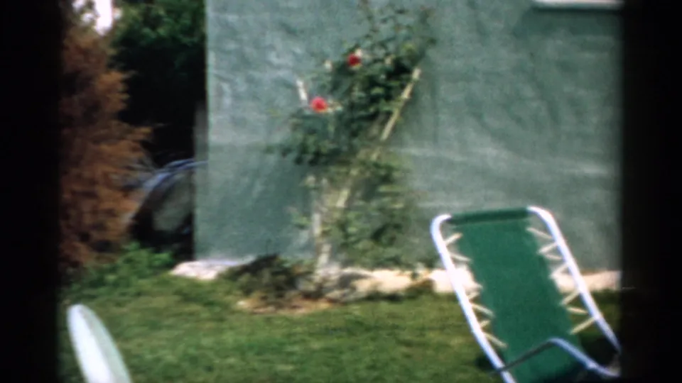 8mm home movie circa 1957 showing a rose trellis with a single red bloom against a stucco garden wall, with a folding green metal lawn chair in the foreground.