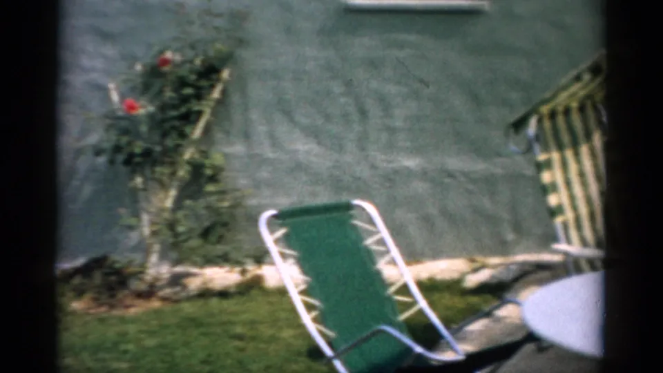 1957 home movie frame: Catskill, NY backyard with two aluminum lawn chairs and a climbing rose bush in bloom against a green-painted exterior wall.