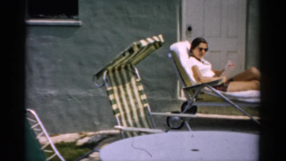 1957 home movie: woman in sunglasses and shorts holds reading material while reclining in a striped aluminum lounge chair in a Catskill, NY backyard.