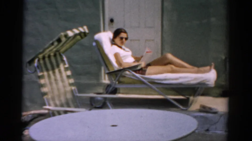 1957 home movie: woman in white top and shorts sits reclined in an aluminum lawn chair, house exterior door visible in the green-painted wall behind, Catskill NY.