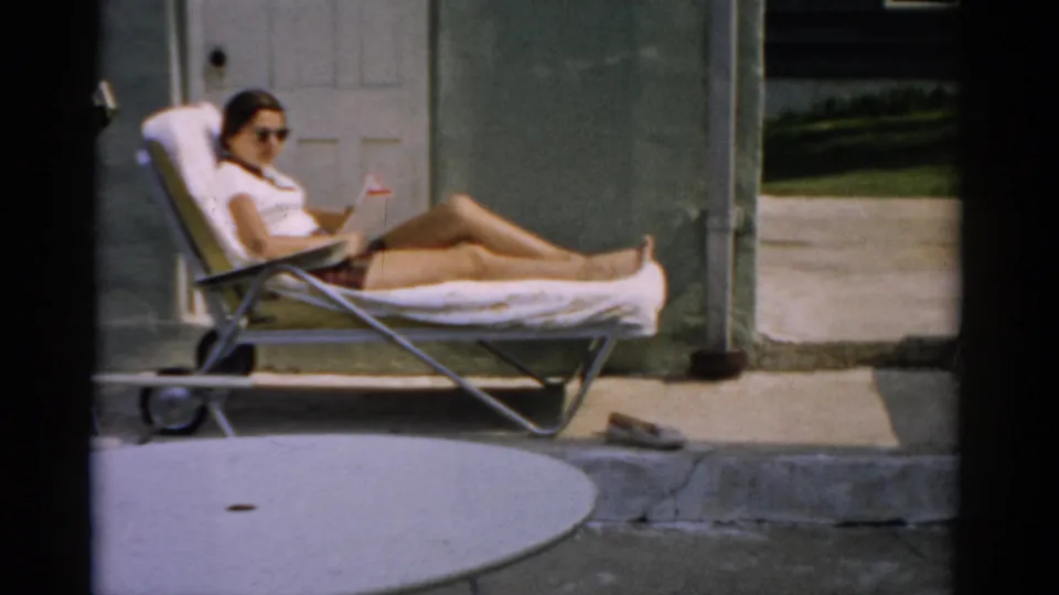 Final 1957 home movie frame: woman relaxing in a green-and-white striped aluminum lounge chair in a Catskill, NY backyard, concrete patio visible in the foreground.