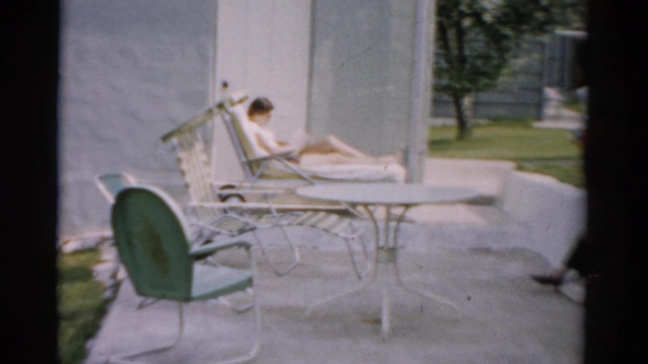 Woman reclining on white metal lounge chair on a concrete patio, reading; mid-century outdoor furniture visible in foreground.