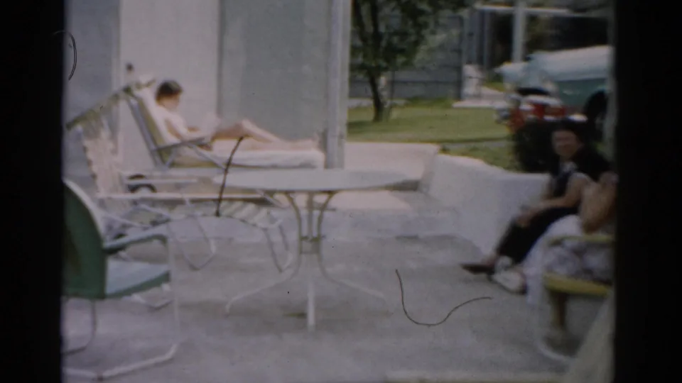 Wider shot of sunlit patio with two women and mid-century outdoor furniture; partial view of a vintage automobile in background, Catskill New York, c. 1957.