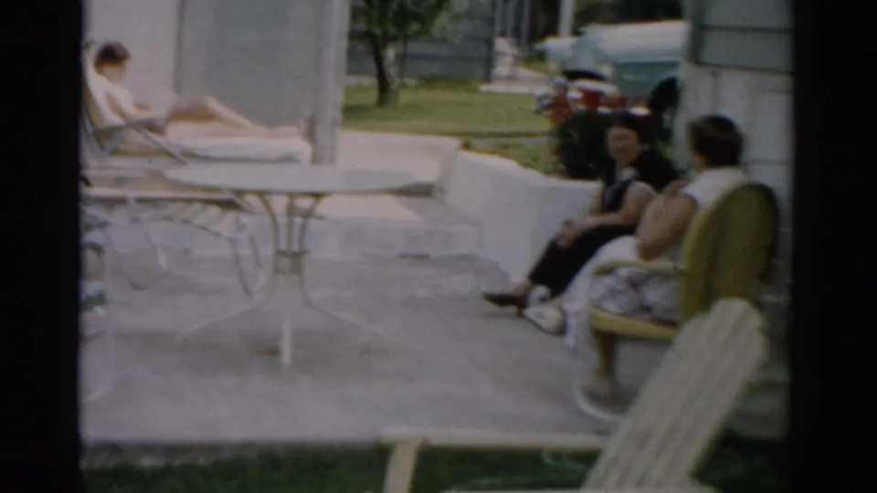 Two women in 1950s dress conversing on patio chairs with a flowering shrub behind them; woman reading in background; Catskill New York area, c. 1957.