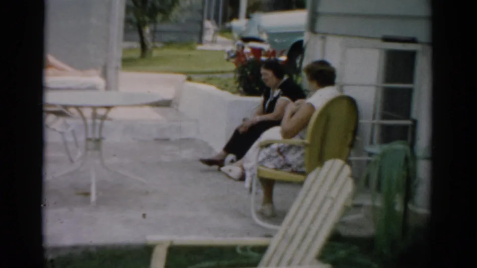 Close view of two women on a sunny patio in mid-century summer dress; residential exterior with window visible at edge; Catskill New York, c. 1957.