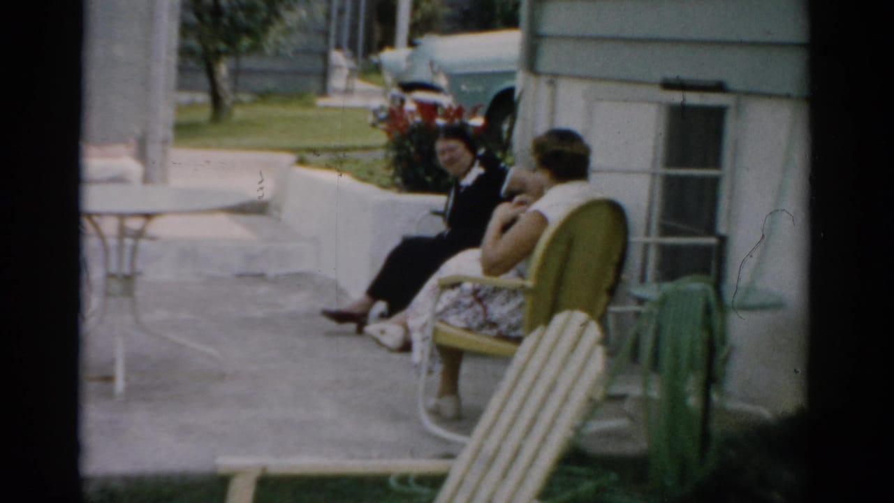 Two women seated in striped metal lawn chairs on a concrete patio beside a house, with a late-1950s car visible in the background.