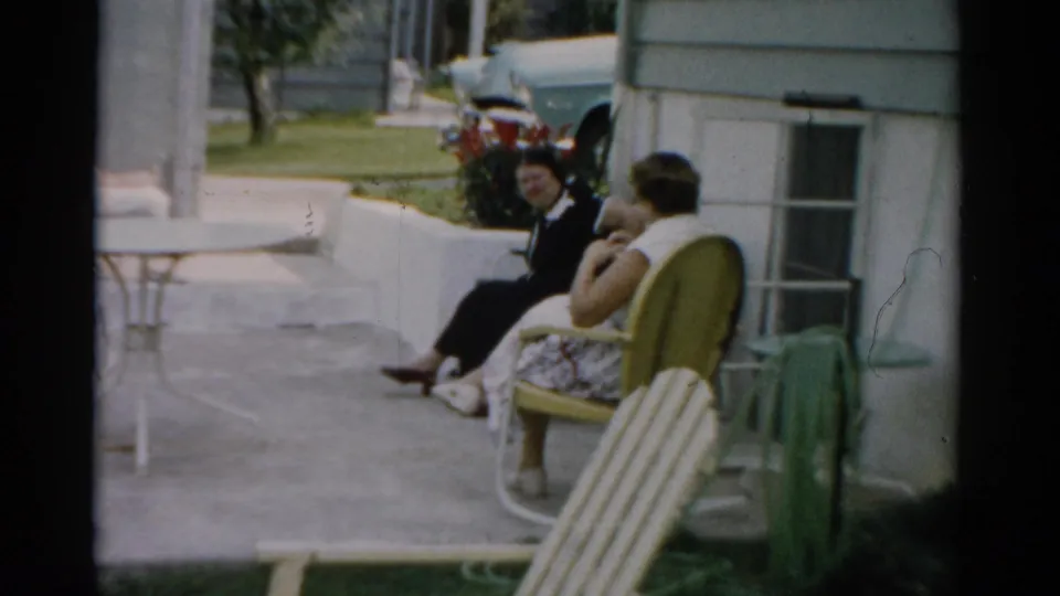 Two women in conversation in striped 1950s lawn chairs on a concrete patio in Catskill, New York, circa 1957; a large blue automobile is parked behind them.