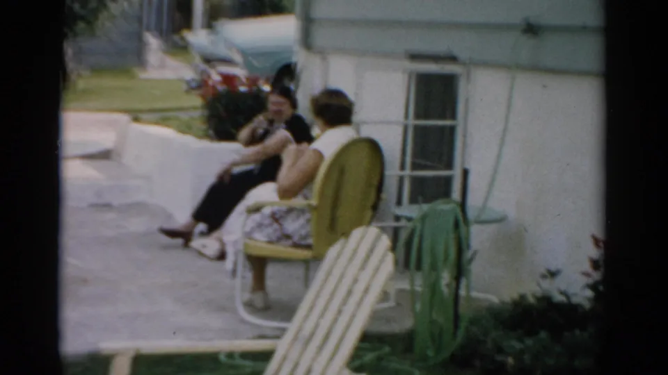 Close view of two women in conversation in striped 1950s lawn chairs on a concrete patio, Catskill, New York, circa 1957; a flowering shrub and car are visible in the background.
