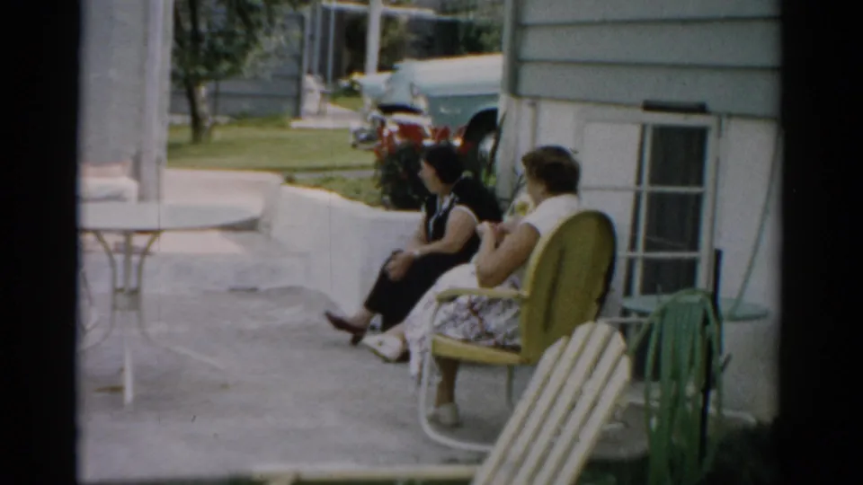 Two women in conversation on a concrete patio in Catskill, New York, circa 1957; a wire patio table and late-1950s automobile are visible in the background.