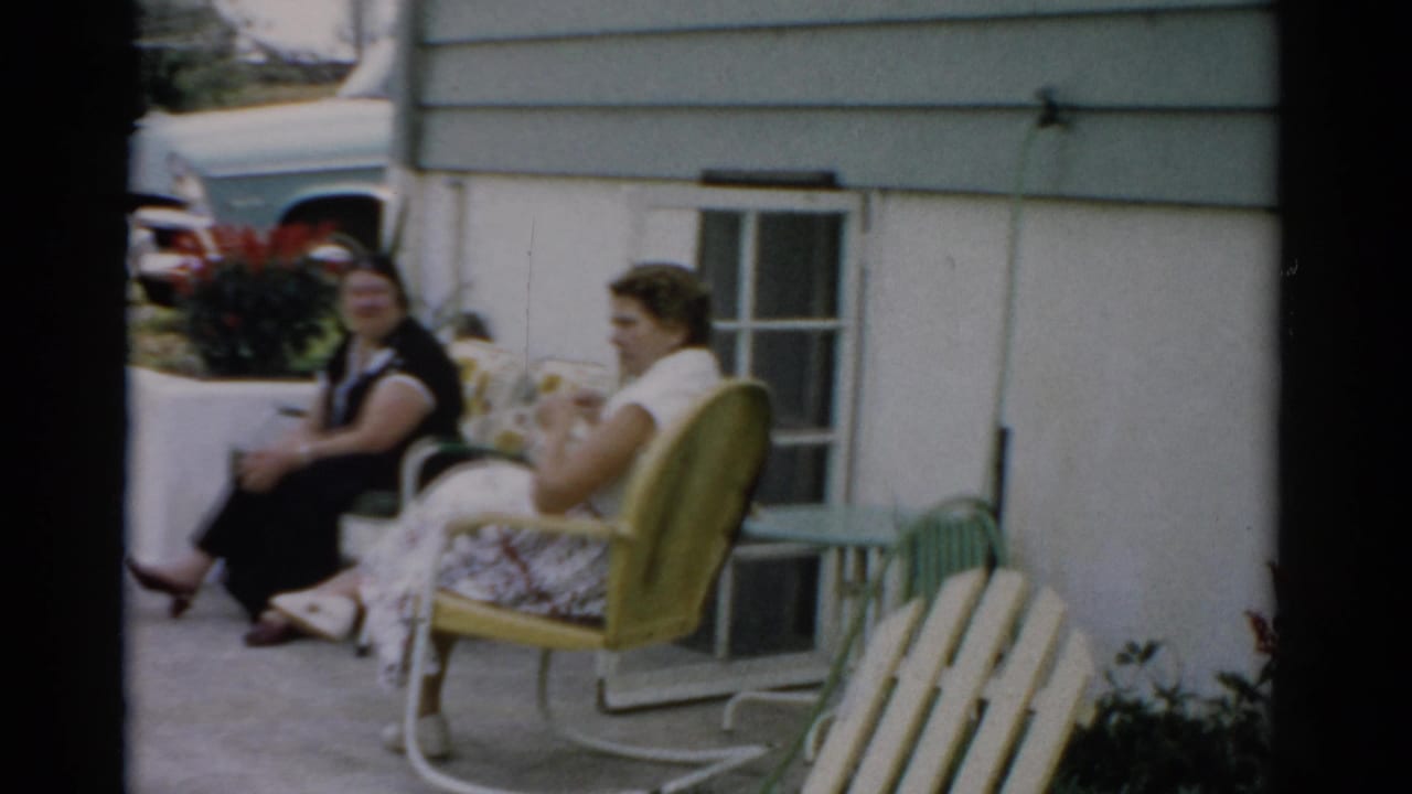 Two women seated outdoors near a gray clapboard house, one in a dark dress and one in a yellow metal lawn chair, in conversation.
