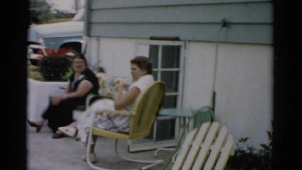 Two women in conversation outside a residential home, circa 1957, Catskill, New York; one seated in a vintage yellow metal lawn chair.