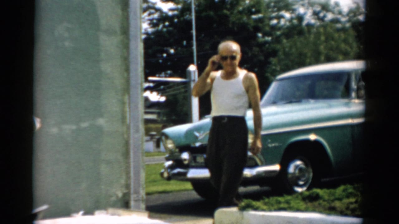 Bald older man in white tank undershirt and sunglasses walks toward camera with hand raised to ear; teal 1950s sedan parked on grass behind him.