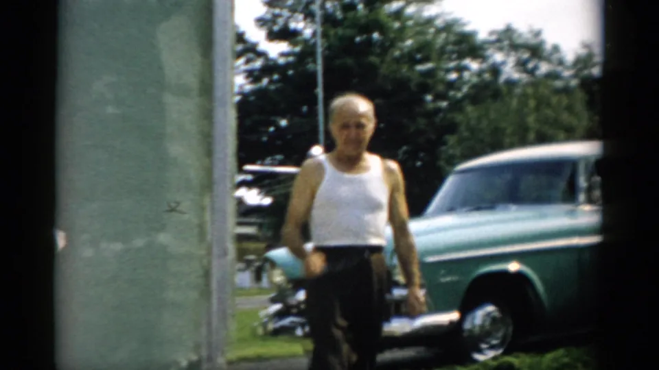 Older man in white ribbed tank undershirt and dark trousers walking toward camera, item held in hand; teal late-1950s American sedan on lawn, circa 1957 Catskill, NY area.