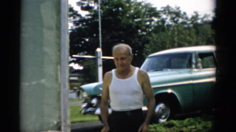 Older man in white ribbed undershirt and dark trousers approaching camera at closer range; teal late-1950s sedan and mature trees behind him, summer setting, circa 1957 Catskill, NY area.