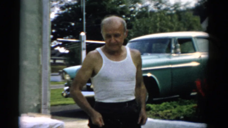 Close shot of older man in white ribbed undershirt looking down; teal 1950s sedan behind him with a second vehicle partially visible at frame right, circa 1957 Catskill, NY area.