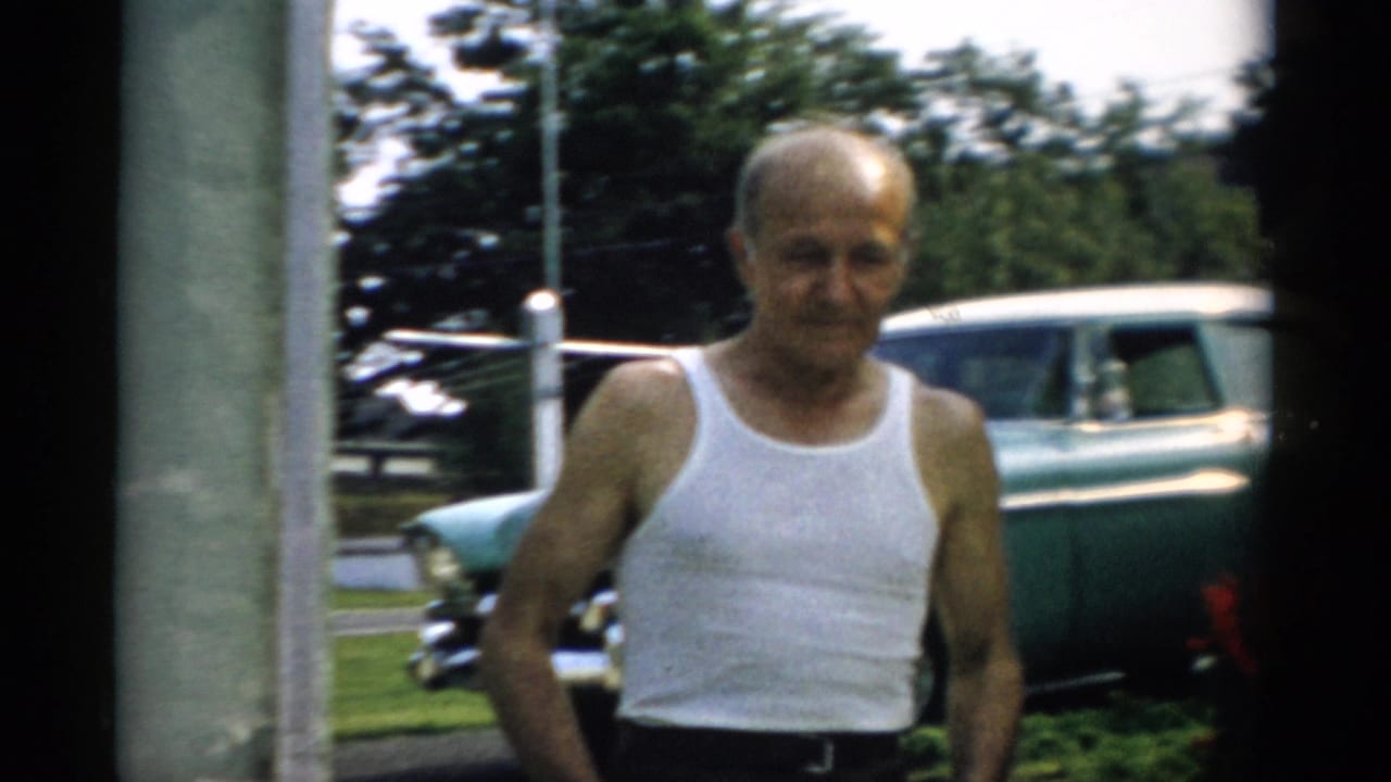 Elderly man in white sleeveless undershirt faces the camera in a Catskill, New York backyard in 1957, a teal mid-century sedan parked in the background.