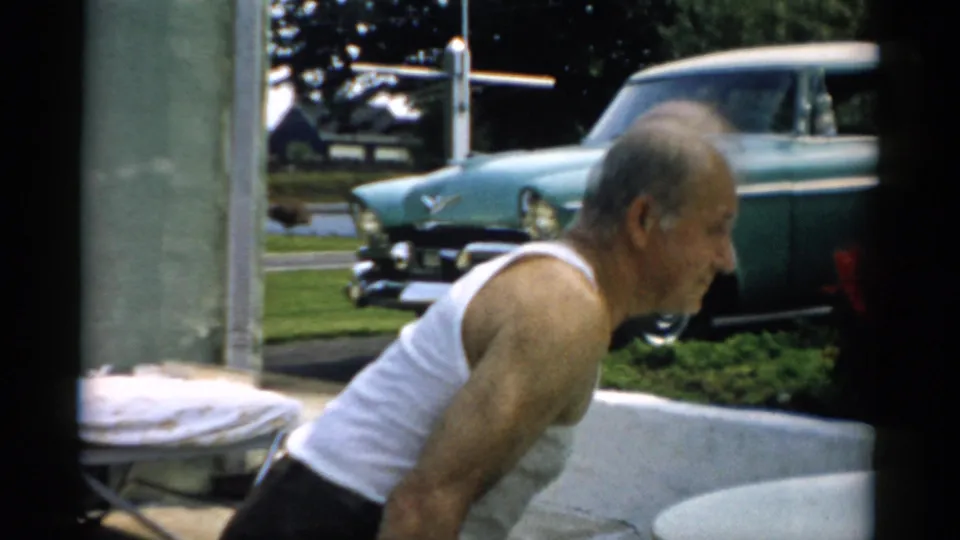 Elderly man turns in profile against a parked teal mid-century American sedan, a covered lounge chair visible at right, Catskill, New York, 1957.