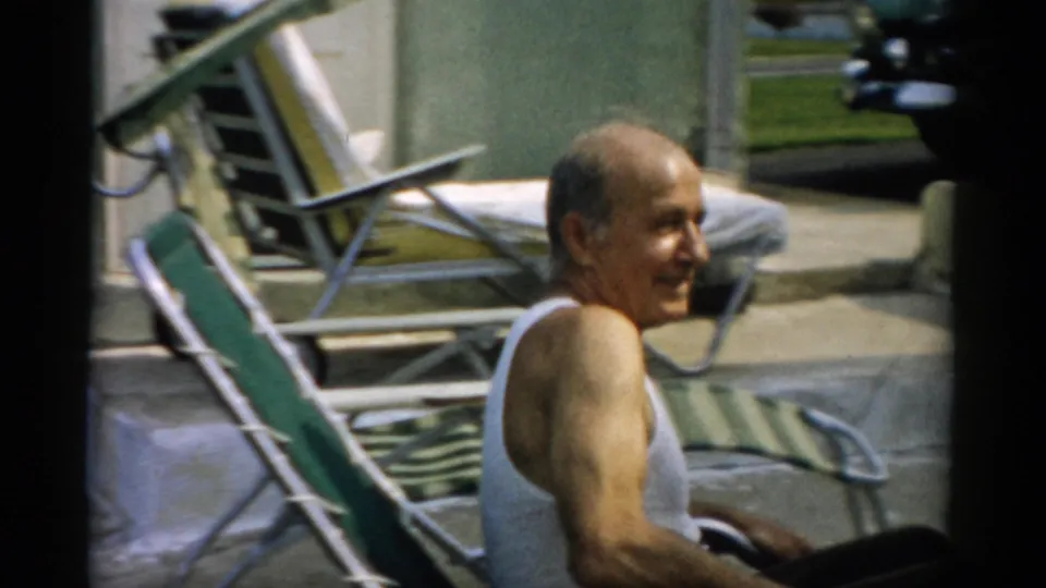 Elderly man settles into a striped aluminum chaise lounge, smiling toward the camera, additional lawn furniture stacked behind him, Catskill, New York, ca. 1957.