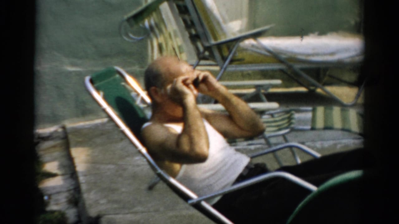Bald man in white tank top adjusting dark sunglasses while seated in a green metal reclining lawn chair outdoors.