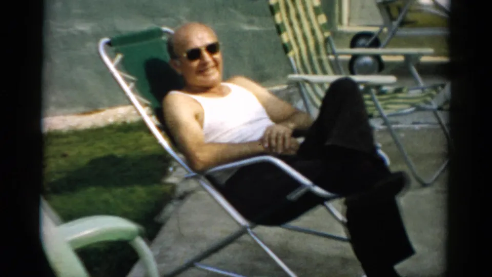 Bald man settled back and smiling toward camera in a green metal reclining lawn chair, 8mm home movie, circa 1957, Catskill, New York.
