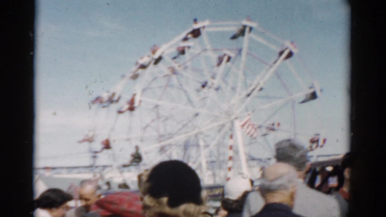 White Ferris wheel above a crowd of fairgoers seen from behind, blue sky, 8mm color film.