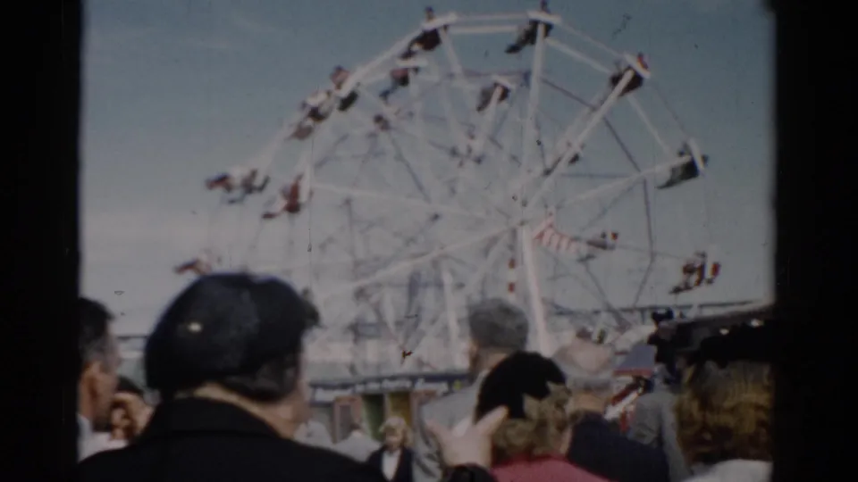 Low-angle view of white Ferris wheel at a 1950s American fair; a fairgoer with a camera visible in the crowd, Catskill area, c. 1957.