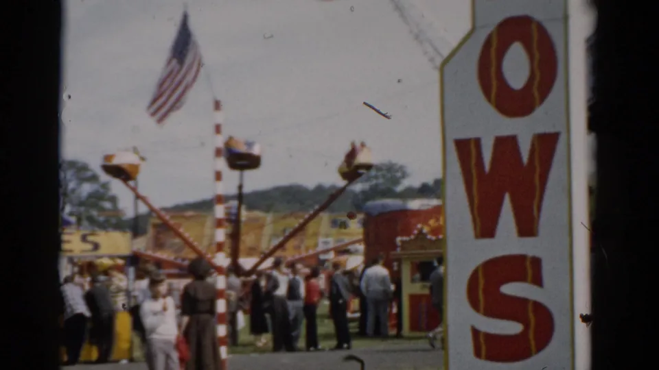 Fairground scene with American flag, partial commercial sign reading 'OWS', a spinning carnival ride, and rolling green hills; Catskill area fair, c. 1957.