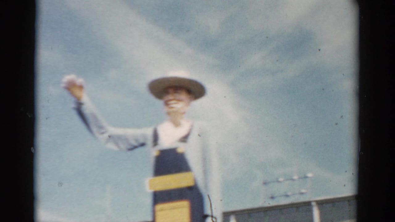 Farmer mascot sculpture at a 1957 New York fairground, wearing blue denim overalls and wide-brimmed hat, one arm raised; 8mm color film.