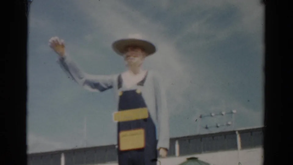Oversized farmer figure at a mid-1950s New York fairground, arm raised, with white fairground building roofline visible in background; 8mm color film.