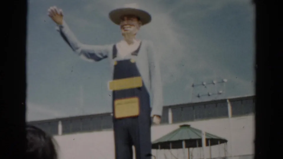 Wider view of the farmer mascot sculpture at a 1957 New York fair, with green-roofed gazebo pavilions and fairgoers visible at bottom of frame.