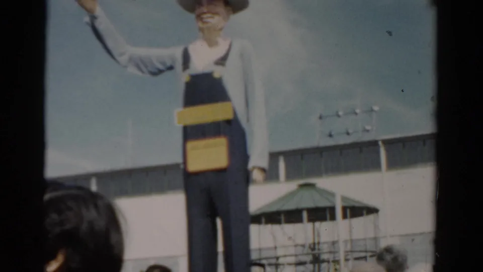 Farmer figure at a mid-century New York fairground, blue overalls and hat, with green pavilion rooftop and fair visitors visible in foreground.
