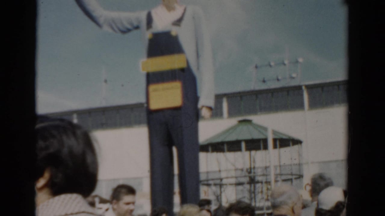 Giant promotional figure in blue denim overalls at a fairground, visitors in 1950s attire below, green gazebo and white building behind, Catskill, New York, circa 1957.