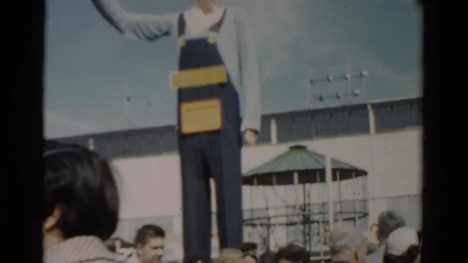Giant promotional figure in blue denim overalls at a fairground, visitors in 1950s attire below, green gazebo and white building behind, Catskill, New York, circa 1957.