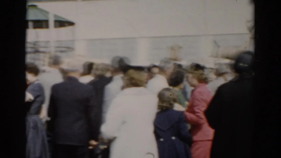 Dense crowd of fairground visitors seen from behind moving through outdoor grounds, white building in background, Catskill, New York, circa 1957.