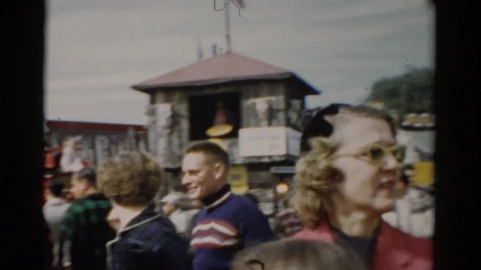 Smiling adult visitors in foreground with stone tower structure with red roof and fairground signage behind, Catskill, New York, circa 1957.