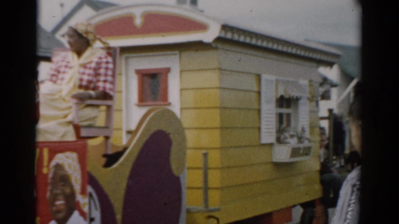 Side view of the yellow-and-maroon Aunt Jemima parade float moving through a crowd, Catskill NY area, c.1957; woman in checkered shirt remains seated on top.