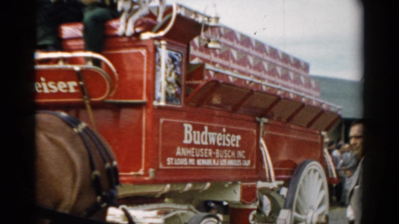 The side of the red Budweiser wagon passes the camera, showing Anheuser-Busch lettering, a large white wheel, harnessed horse parts, and adults near the street edge.