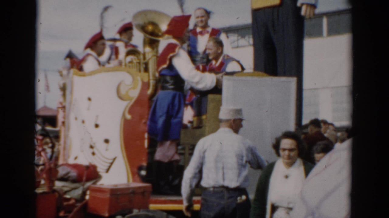 Band members in red and blue costumes on a decorated wagon unloading a large brass instrument; spectators watch from ground level.
