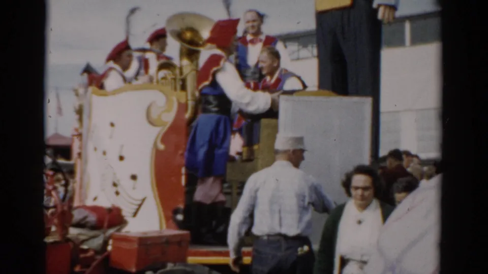 Band members in ornate red-and-blue costumes handle a brass tuba on a decorated fair wagon while civilian spectators in 1950s dress look on, Catskill, New York, 1957.