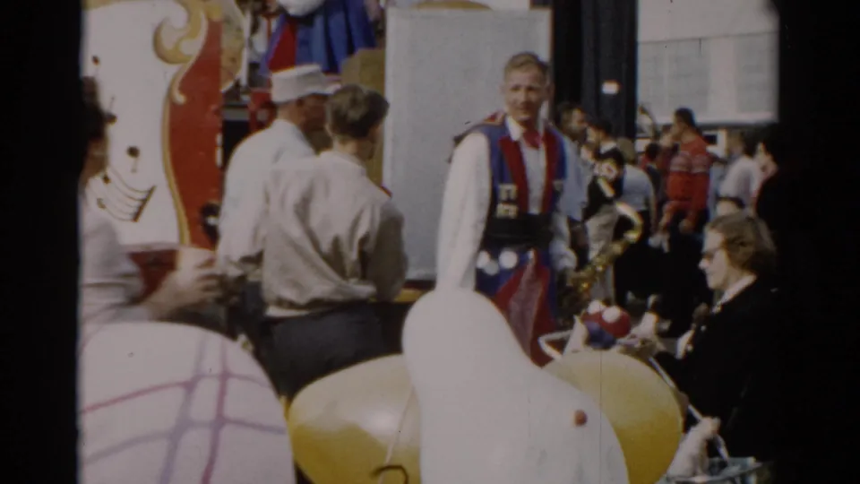 A band member in red-and-blue ceremonial costume faces the camera near an ornate fair wagon, surrounded by 1950s-dressed spectators; yellow props visible at right, Catskill, New York, 1957.
