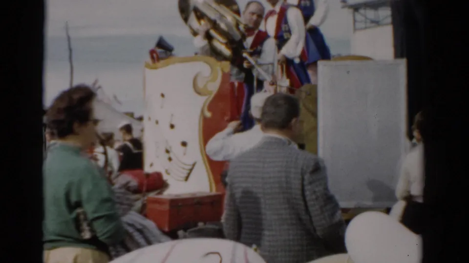Ground-level view of spectators and costumed band members around a red-and-gold ornamental fair wagon; woman in green jacket in foreground, Catskill, New York, 1957.
