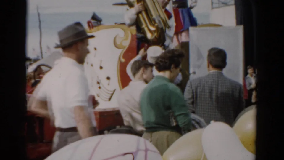 Man in a gray felt hat and white shirt stands prominent in the foreground crowd near the decorated fair wagon; yellow round props or balloons visible at bottom, Catskill, New York, 1957.