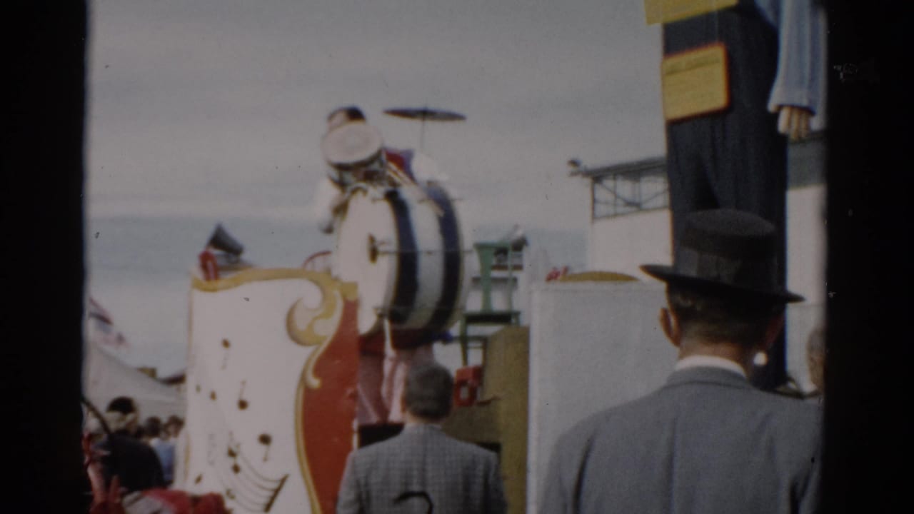 Decorated parade float with large bass drum viewed from behind a crowd of adults at a 1950s fair.